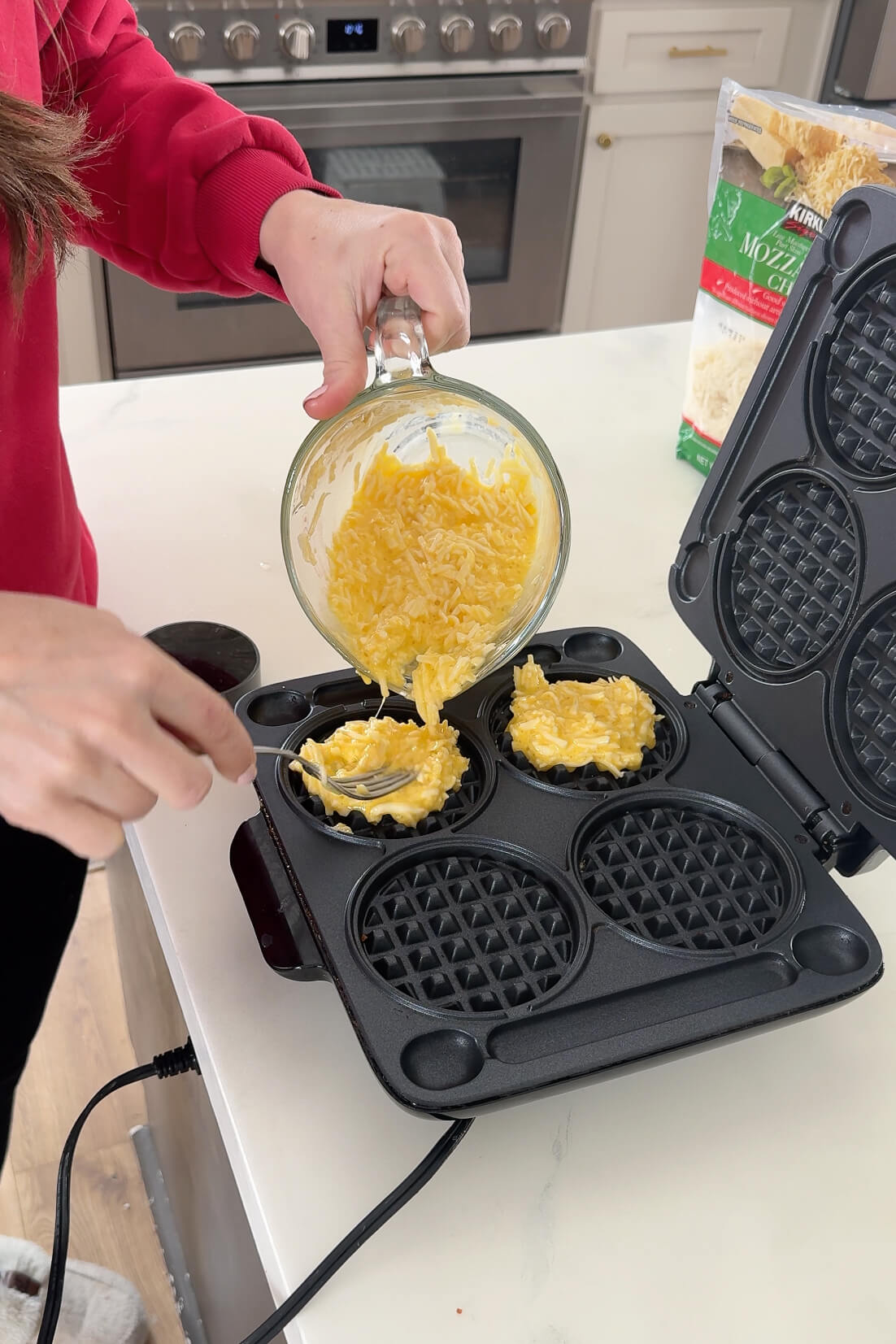 A woman in a red sweater pours a cheesy egg mixture into the circular wells of an open waffle maker, preparing chaffles. The kitchen background features a stove and a bag of shredded mozzarella cheese.